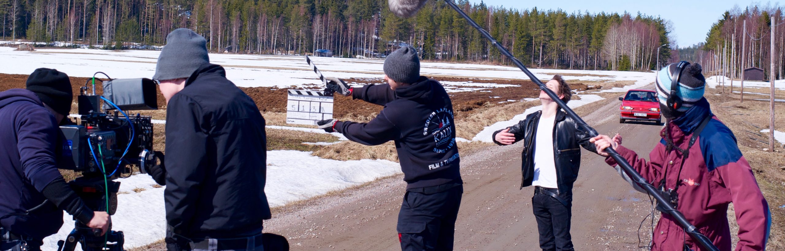 Filminspelning, filmkameran är riktad mot en klappa, i bakgrunden står en man på en landsväg och en röd bil kommer åkandes mot honom.